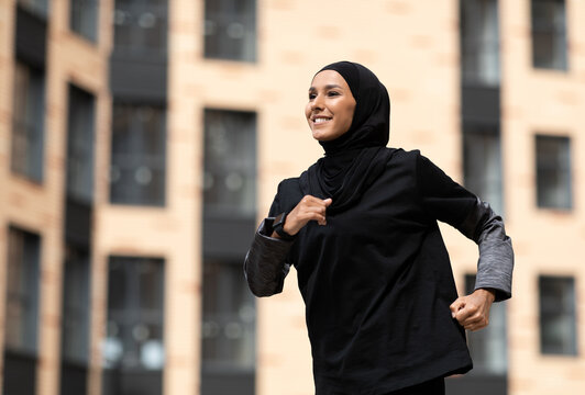 Glad smiling young arab female in hijab with fitness tracker running on building background outdoor. Fitness outside, jogging in morning, body and health care, training, ad and great sports offer - Powered by Adobe