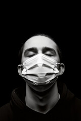 Fototapeta na wymiar Young man wears a surgical mask and closes his eyes. The person is dressed in a hoodie and photographed against a dark black background in monochrome. Quarantine and pandemic concept. Generative AI.
