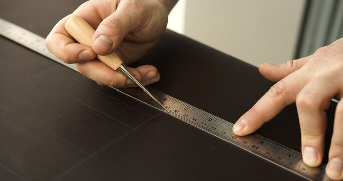 Hands of craftsman marking straight lines on leather sheet with awl and metal ruler for leatherwork