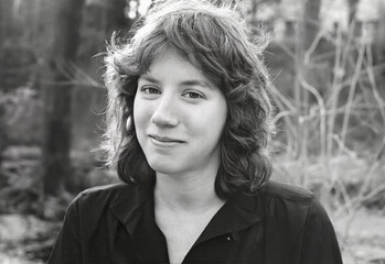 Black and white film portrait of a young woman with natural expression, standing outdoors with soft backlight and visible film grain texture.
