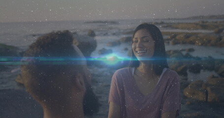 Sitting Hispanic couple wearing purple T-shirt and casual top on rocky shore at sunset, copy space