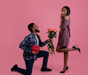 Passionate black man giving his sweetheart bouquet of roses and heart shaped gift for Valentine's Day on pink studio background. Happy woman getting flowers and present from her boyfriend, full length