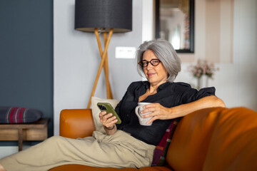 Senior woman relaxing on sofa with smartphone and coffee