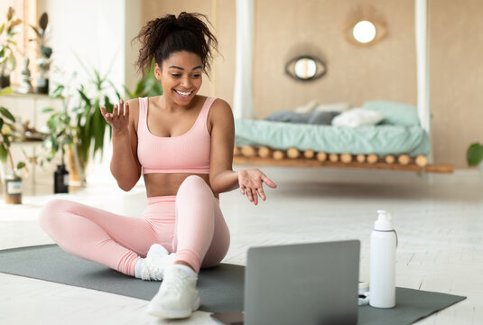 Online exercise session marathon. Happy black woman sitting on floor mat and talking with coach, using pc for virtual workout, exercising at home, free space