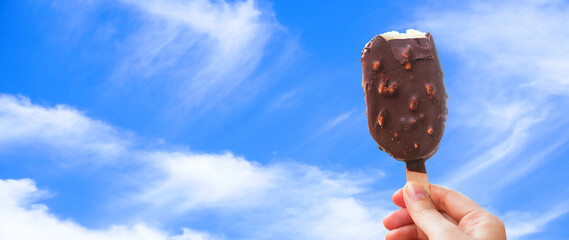 Chocolate covered ice cream bar with blue sky and cloud summer at the beach, during the holidays. Hand holds delicious chocolate and nuts almond ice cream bar on a stick.