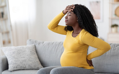 African american pregnant woman suffering from headache at home, sitting on couch, touching her...