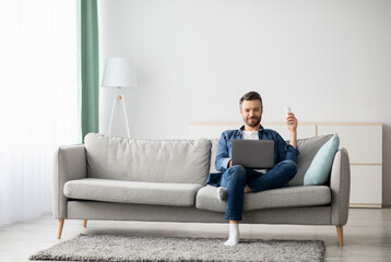 Obraz premium Relaxed middle-aged man with laptop sitting on couch at home, surfing on Internet or watching movie, raising hand with AC remote up, copy space. Cooling systems for home, air conditioner concept