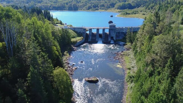 Panoramic aerial view looking down towards a lake and an old hydroelectric dam, passing inside its concrete structure. Mitis Dam, Lake & Mitis River, Quebec, Canada, 2025.