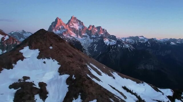 Aerial view of snow capped mountains with rocky peaks and clear blue sky at golden hour light