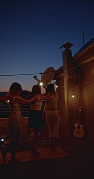 Group of diverse young friends joyfully dancing and celebrating together on a rooftop party at twilight. Enjoying a fun, casual social gathering with illuminated string lights and city views.