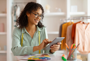 Cheerful fashion designer using a digital tablet beside a color chart in a well-organized creative studio, embodying professional creativity and modern entrepreneurship