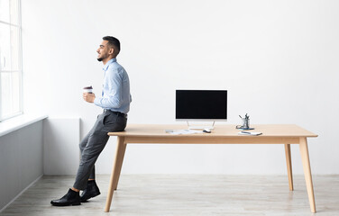 Smiling Successful Bearded Man In Formalwear Sitting Leaning On Table, Drinking Coffee Looking At Window, Showing Pc With Blank Black Mock Up Screen At Home Office, Free Copy Space, Full Body Length © Prostock-studio