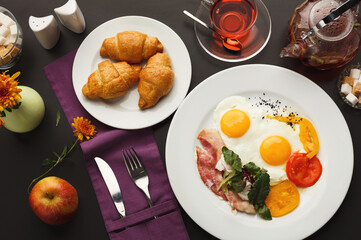 Restaurant delicious breakfast with sunny side up fried eggs, bacon, vegetables and greens, croissants and freshly brewed tea served on black table. Nourishing morning meals, top view