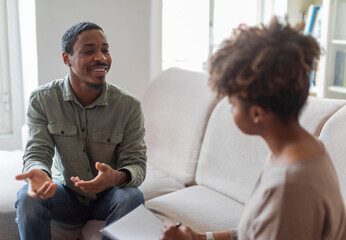 Cheerful positive handsome young african american guy in casual outfit sitting on couch at cozy counselor office, having conversation with therapist black woman, sharing good results of therapy
