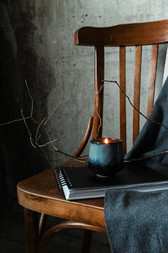 Lit candle in ceramic cup on notebook with wooden chair and gray fabric. Cozy minimal still life in wabi-sabi mood.