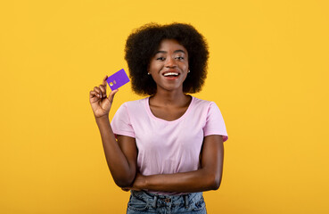Easy payment concept. Happy young black woman holding debit credit card in hand, smiling and showing it to camera, posing isolated over yellow studio background