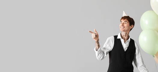 Young tattooed man with balloons celebrating Birthday and pointing at something on grey background with space for text