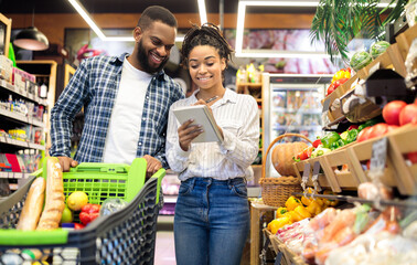 Grocery Shopping. Black Family Couple Buying Food And Fruits Taking Notes In Checklist Choosing Groceries Together In Modern Supermarket. Spouses Buy Fresh Organic Vegetables In Store