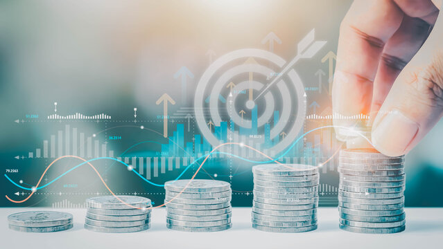 A hand stacks coins on a growing pile before a digital interface with charts and a target. This represents financial planning, savings, investment growth, and achieving monetary objectives.