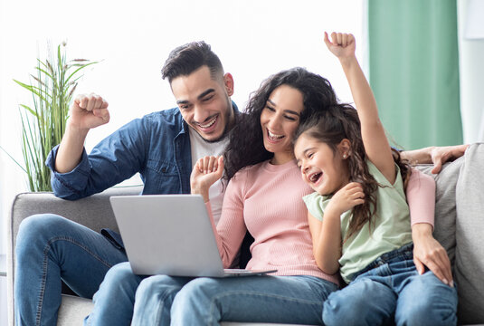 Portraif of happy arabic family of three celebrating success with laptop at home, cheerful middle eastern mother, father and daughter looking at computer screen and raising hands with excitement
