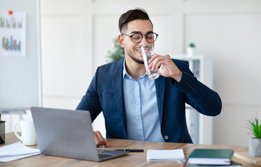 Cool Arab businessman in suit drinking water while working on laptop at modern office. Millennial...