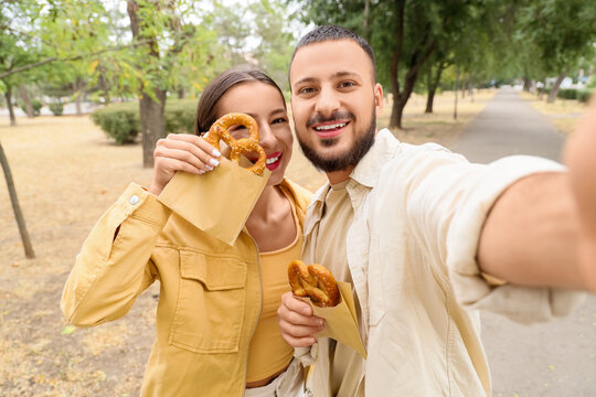 Young happy couple with tasty pretzels taking selfie while walking in park - Powered by Adobe