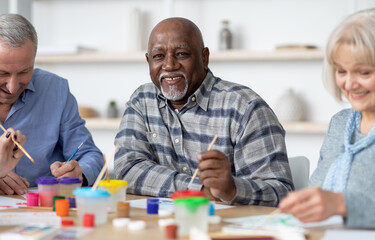 Leisure for senior people at retirement house concept. Happy senior african american man enjoying painting activity with his multiracial friends, holding brush and smiling at camera