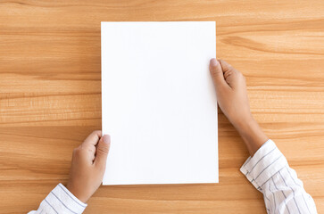 Overhead view of woman holding blank paper sheet A4 size or letter paper on wooden desk background,...