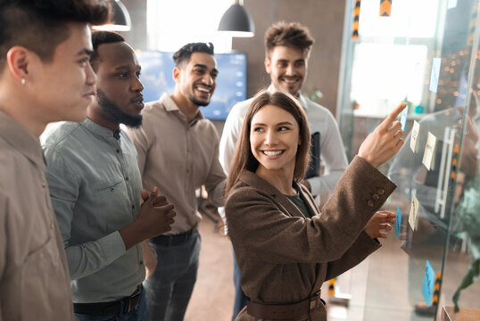 Teamwork Concept. Creative group of smiling professional businesspeople brainstorming, wonan pointing at sticky post-it paper notes on glass wall, discussing ideas analysing new business strategy