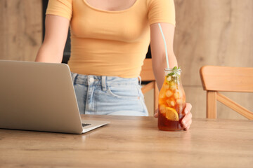 Beautiful young woman with laptop and bottle of tasty cold tea on table in kitchen, closeup