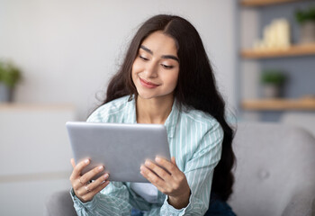 Charming Indian lady using tablet computer, studying remotely or having online conference at home. Young woman with touch pad taking part in web meeting or watching movie indoors