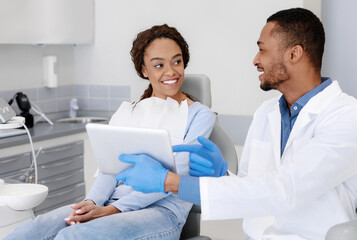 Fototapeta premium Cheery african guy dentist showing woman patient his clinic website on digital tablet