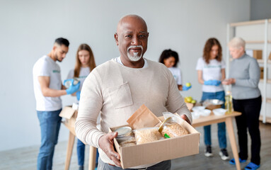 African american senior man holding box with donations food, looking and smiling at camera while group of volunteers packing boxes with products for poor and homeless people