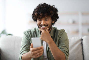 Cheerful curly millennial hindu guy in casual sitting on couch at home, using brand new cell phone...