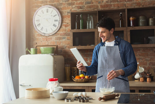 Serious young man baking pie in loft kitchen at home, using digital tablet with recipe, copy space - Powered by Adobe