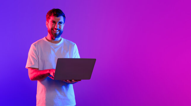 Young caucasian guy using laptop standing in studio with purple and blue neon light, showcasing concept of online work and gaming. Technology and freelance. Panorama, copy space