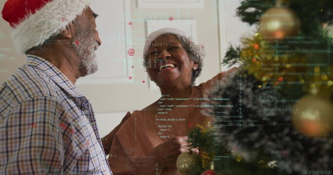 Smiling couple wearing Santa hats, holding bauble by Christmas tree at home, with code overlay