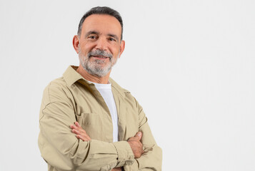 Fototapeta premium Handsome Senior Man Posing With Folded Arms Over White Studio Background, Portrait Of Confident Elderly Gentleman Wearing Beige Casual Shirt, Smiling And Looking At Camera, Copy Space