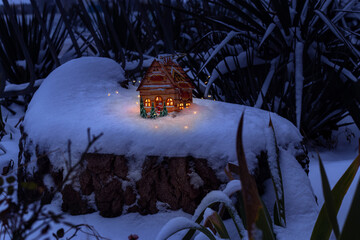 Christmas lantern in form of house standson snow on large stump in winter garden. Windows of house are burning with bright light, Christmas tree garland lighting. 