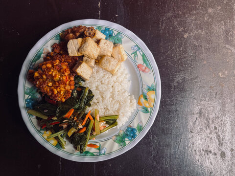 Top view of Indonesian Food with side dishes of mix vegetables, tofu, frikadel or corn fritters, fried chicken, and steamed rice as main food on white plate. Isolated by brown wooden