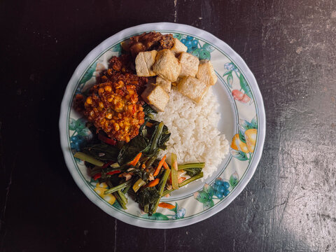 Top view of Indonesian Food with side dishes of mix vegetables, tofu, frikadel or corn fritters, fried chicken, and steamed rice as main food on white plate. Isolated by brown wooden