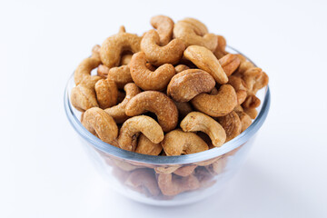 A transparent glass bowl filled with cashews on a white background. The cashews are whole and have a golden-brown hue.
