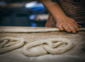 Hands shaping pretzel dough on a floured wooden table. Twisting and forming traditional pretzels before baking, rustic homemade style and artisan preparation.
