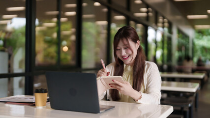 Fototapeta premium Adult Asian freelance professional works productive outdoor cafe writing notes laptop computer natural light happy lifestyle