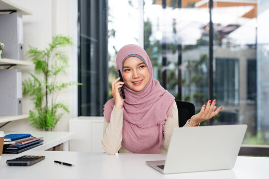 Happy smiling Muslim business woman in hijab professional lady executive manager talking on the phone making business call on cellphone at work in office and using laptop computer. - Powered by Adobe