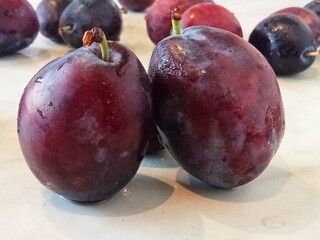 Several ripe plums on a light surface. Two plums are in the foreground, one of them is visible whole, and the other is cut in half, showing the stone and golden flesh.