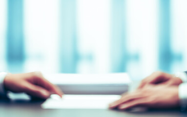 Two people sit at a desk with paperwork and a blurred background.