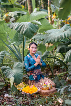 Balinese woman in traditional attire kneeling in prayer beside colorful offerings, surrounded by lush tropical leaves.