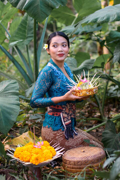 Woman in blue kebaya sitting in jungle with offerings