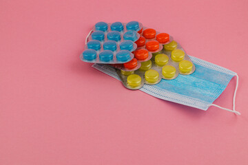Colorful medication capsules and face masks arranged on a pink background